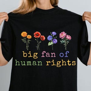 May include: Black t-shirt with a floral design and the text "big fan of human rights" in rainbow colors. The shirt features illustrations of orange, red, blue, and pink flowers.