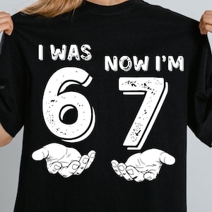 May include: Black t-shirt with the text "I WAS 67 NOW I'M" in white. The numbers 6 and 7 are large and white, with a distressed texture. The number 6 is above a pair of open hands.