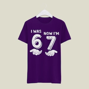 May include: A purple t-shirt with the text "I WAS NOW I'M 67" in white. The number 67 is large and stylized, with open hands below. The shirt is hanging on a white hanger.