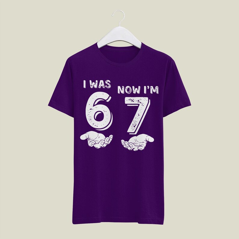 May include: A purple t-shirt with the text "I WAS NOW I'M 67" in white. The number 67 is large and stylized, with open hands below. The shirt is hanging on a white hanger.