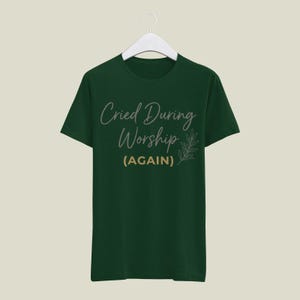 May include: A dark green t-shirt with the text "Cried During Worship (AGAIN)" in a script font. A small leaf design is to the right of the text. The shirt is hanging on a white hanger.
