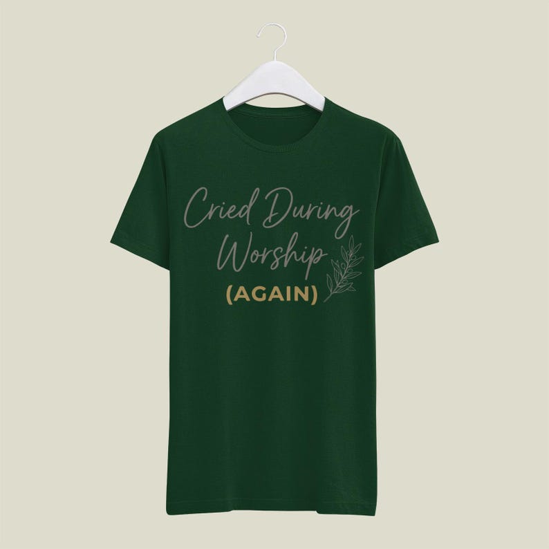 May include: A dark green t-shirt with the text "Cried During Worship (AGAIN)" in a script font. A small leaf design is to the right of the text. The shirt is hanging on a white hanger.