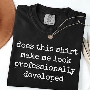 May include: A black short-sleeved t-shirt with a crew neckline and a Comfort Colors brand label. The shirt features white text printed on the front that reads, does this shirt make me look professionally developed, in a typewriter-style font.