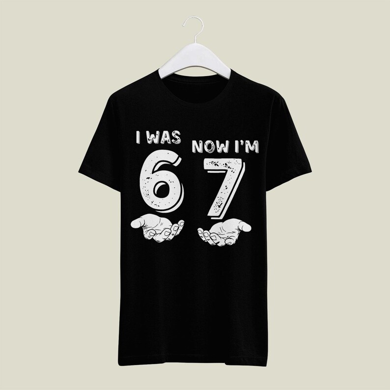 May include: Black t-shirt with the text "I WAS NOW I'M 67" in white, with the number 6 and 7 held up by two hands. The shirt is hanging on a white hanger.