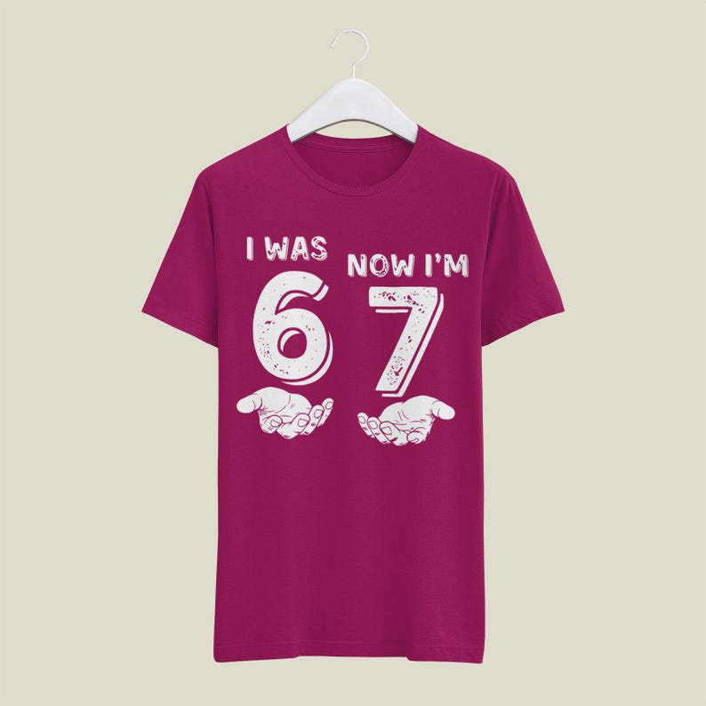 May include: A maroon t-shirt with the text "I WAS NOW I'M 67" in white, distressed font. The number 6 and 7 are large and stylized, with open hands below each number. A fun birthday gift.