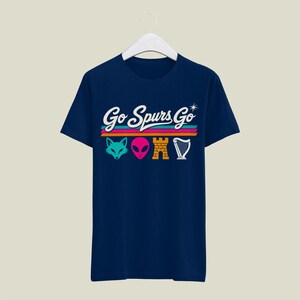 K&ouml;nnte beinhalten: Marineblaues T-Shirt mit dem Text "Go Spurs Go" in wei&szlig;er Schrift &uuml;ber einem horizontalen Streifendesign in T&uuml;rkis, Orange und Pink. Unter den Streifen befinden sich t&uuml;rkisfarbene Fuchs-, rosa Alien-, goldene Burg- und wei&szlig;e Harfengrafiken.