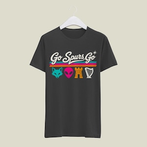 K&ouml;nnte beinhalten: Dunkelgraues T-Shirt mit dem Text "Go Spurs Go" in wei&szlig;er Schrift &uuml;ber einem t&uuml;rkisfarbenen Fuchs, einem rosa Alien, einer orangefarbenen Burg und einer wei&szlig;en Harfe. Der Text und die Grafiken befinden sich &uuml;ber horizontalen Streifen in T&uuml;rkis, Orange und Pink.