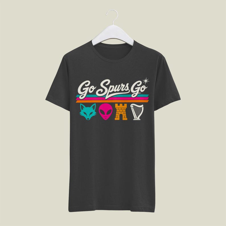 K&ouml;nnte beinhalten: Dunkelgraues T-Shirt mit dem Text "Go Spurs Go" in wei&szlig;er Schrift &uuml;ber einem t&uuml;rkisfarbenen Fuchs, einem rosa Alien, einer orangefarbenen Burg und einer wei&szlig;en Harfe. Der Text und die Grafiken befinden sich &uuml;ber horizontalen Streifen in T&uuml;rkis, Orange und Pink.