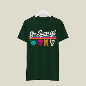 K&ouml;nnte beinhalten: Dunkelgr&uuml;nes T-Shirt mit dem Text "Go Spurs Go" in wei&szlig;er Schrift &uuml;ber einem horizontalen Streifendesign in Pink, T&uuml;rkis und Orange. Darunter befinden sich vier grafische Symbole: ein t&uuml;rkisfarbener Fuchskopf, ein pinkfarbener Alienkopf, ein goldenes Schloss und eine wei&szlig;e Harfe.