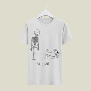 Puede incluir: Una camiseta gris claro con un dibujo lineal en negro de un esqueleto al que le falta una pierna y un perro que sale corriendo con un hueso en la boca. Debajo de la ilustraci&oacute;n, el texto WELL, SHIT... aparece en una fuente negra sencilla.