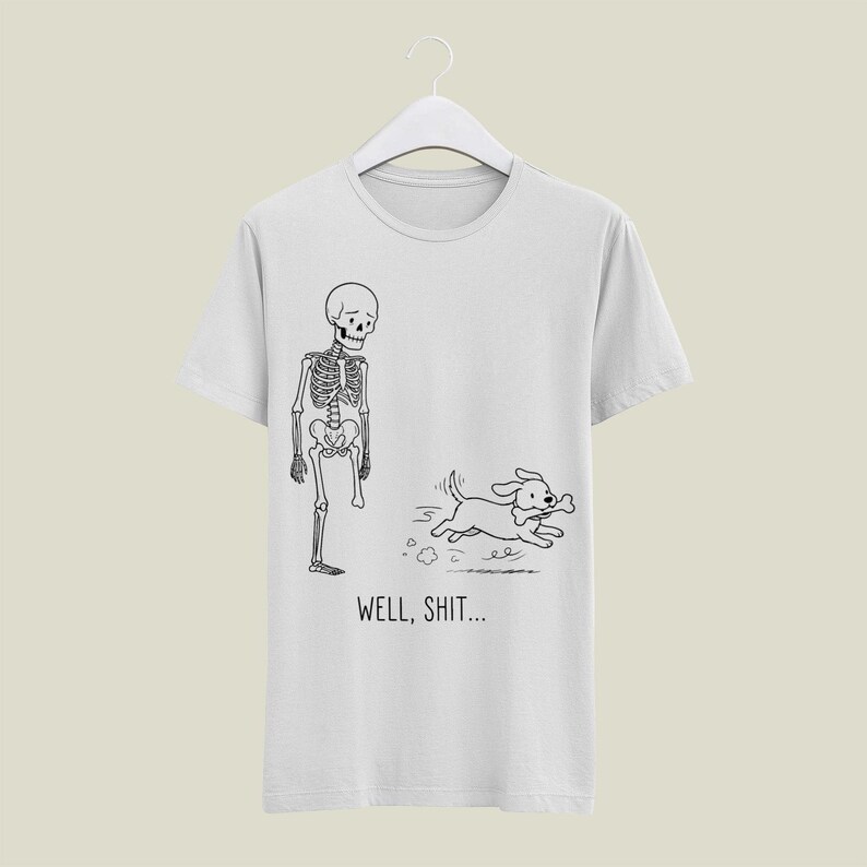 Puede incluir: Una camiseta gris claro con un dibujo lineal en negro de un esqueleto al que le falta una pierna y un perro que sale corriendo con un hueso en la boca. Debajo de la ilustraci&oacute;n, el texto WELL, SHIT... aparece en una fuente negra sencilla.