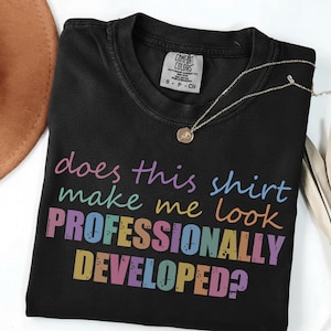 May include: A black crew neck t-shirt featuring the multi-colored text "does this shirt make me look PROFESSIONALLY DEVELOPED?" printed on the front. The shirt has a Comfort Colors brand label on the inside collar indicating a size small.