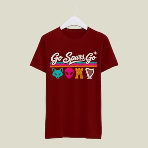 K&ouml;nnte beinhalten: Burgunderfarbenes T-Shirt mit dem Schriftzug "Go Spurs Go" in wei&szlig;er Schrift &uuml;ber einem horizontalen Streifendesign. Unter den Streifen befinden sich vier farbenfrohe grafische Symbole: ein Fuchs, ein Alien, eine Burg und eine Harfe.