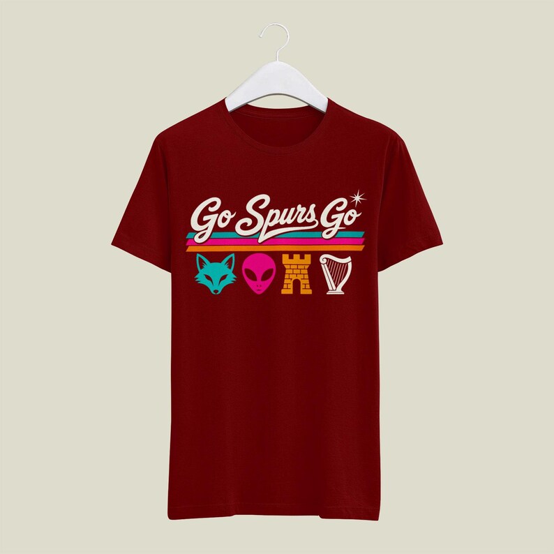 K&ouml;nnte beinhalten: Burgunderfarbenes T-Shirt mit dem Schriftzug "Go Spurs Go" in wei&szlig;er Schrift &uuml;ber einem horizontalen Streifendesign. Unter den Streifen befinden sich vier farbenfrohe grafische Symbole: ein Fuchs, ein Alien, eine Burg und eine Harfe.