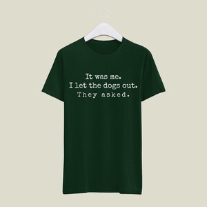 May include: A dark green t-shirt with the text "It was me. I let the dogs out. They asked." in a white, typewriter-style font. The shirt is hanging on a white hanger against a neutral background.