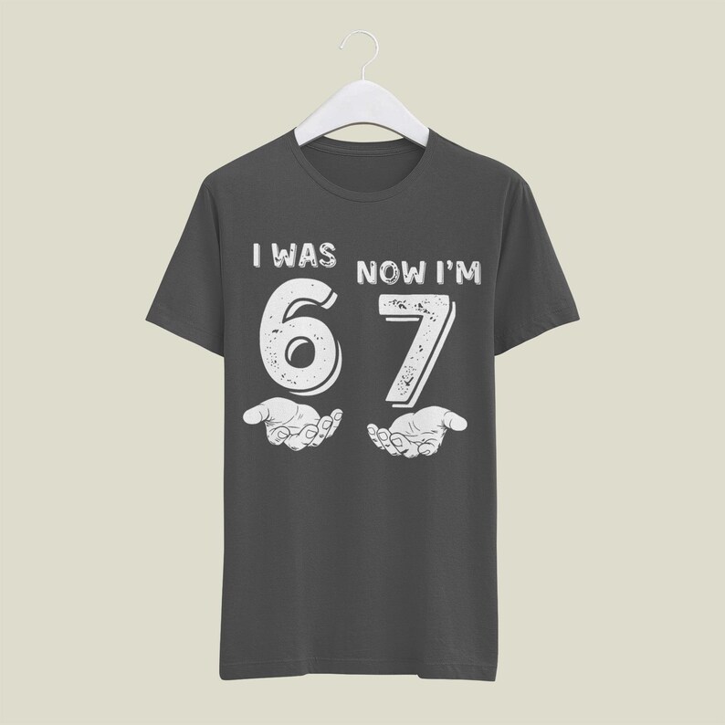 May include: Dark gray t-shirt with the text "I WAS NOW I'M 67" in white, with the numbers 6 and 7 designed to look like they are being held by two hands. The shirt is hanging on a white hanger.