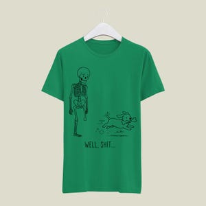 Puede incluir: Una camiseta verde de manga corta con un dibujo lineal negro de un esqueleto al que le falta una pierna y un perro que huye con un hueso en la boca. Debajo de la ilustraci&oacute;n, el texto dice WELL, SHIT... en una fuente negra y sencilla.