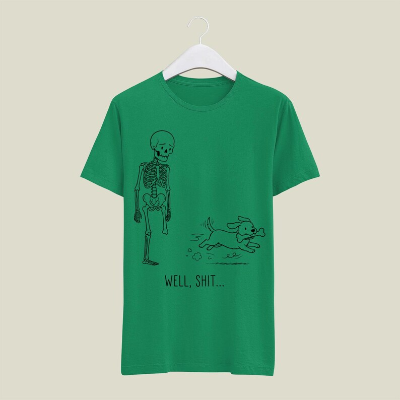 Puede incluir: Una camiseta verde de manga corta con un dibujo lineal negro de un esqueleto al que le falta una pierna y un perro que huye con un hueso en la boca. Debajo de la ilustraci&oacute;n, el texto dice WELL, SHIT... en una fuente negra y sencilla.