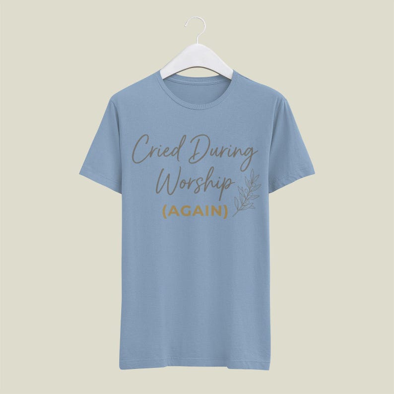 May include: A light blue t-shirt with the text "Cried During Worship (AGAIN)" in a script font. A leaf design is to the right of the text. The shirt is hanging on a white hanger.