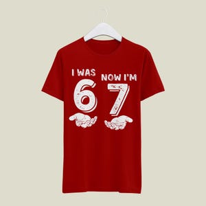 May include: Red t-shirt with the text "I WAS NOW I'M 67" in white, distressed font. The number 67 is large and centered, with stylized hands below. The shirt is hanging on a white hanger.
