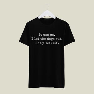 May include: Black t-shirt with the text "It was me. I let the dogs out. They asked." in a white, typewriter-style font. The shirt is hanging on a white hanger against a neutral background.