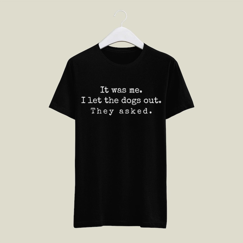 May include: Black t-shirt with the text "It was me. I let the dogs out. They asked." in a white, typewriter-style font. The shirt is hanging on a white hanger against a neutral background.