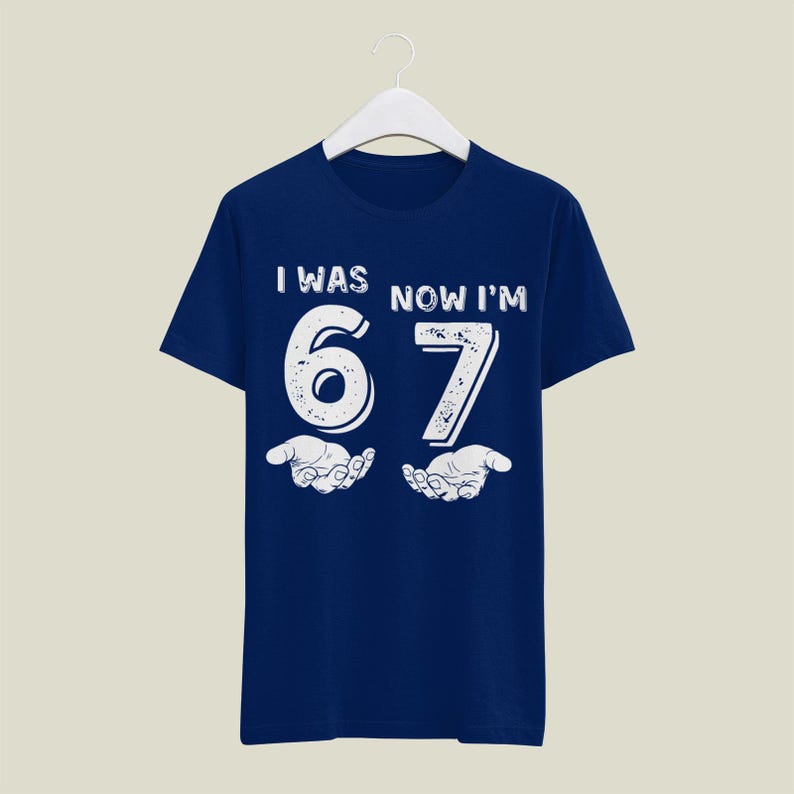 May include: Navy blue t-shirt with the text "I WAS NOW I'M 67" in white, distressed font. The number 6 and 7 are large and stylized, with open hands beneath. A fun, birthday-themed graphic tee.