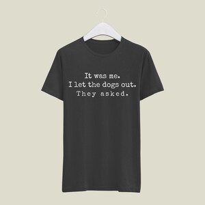 May include: Black t-shirt with the text "It was me. I let the dogs out. They asked." in a white, typewriter-style font. The shirt is hanging on a white hanger against a neutral background.