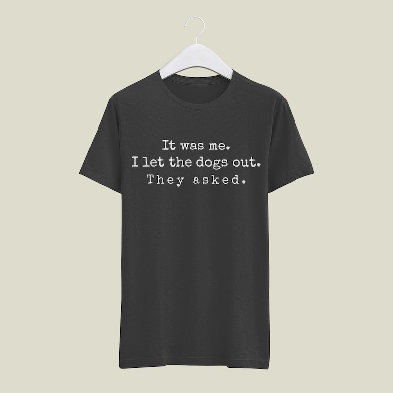 May include: Black t-shirt with the text "It was me. I let the dogs out. They asked." in a white, typewriter-style font. The shirt is hanging on a white hanger against a neutral background.