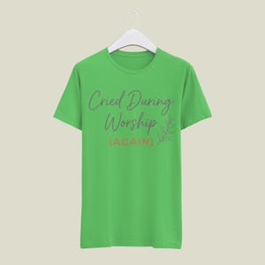 May include: A lime green t-shirt with the text "Cried During Worship (AGAIN)" in a script font. A small leaf design is to the right of the text. The shirt is hanging on a white hanger.
