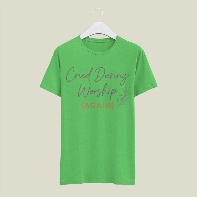 May include: A lime green t-shirt with the text "Cried During Worship (AGAIN)" in a script font. A small leaf design is to the right of the text. The shirt is hanging on a white hanger.