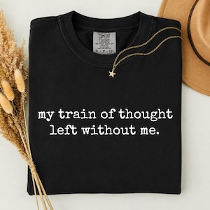 Op de afbeelding: Een zwart T-shirt met korte mouwen en een ronde hals, voorzien van de witte tekst "my train of thought left without me." op de voorzijde. Het shirt heeft een Comfort Colors merklabel in de kraag en een gouden ketting met een ster.