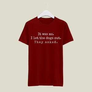 May include: A maroon t-shirt with the text "It was me. I let the dogs out. They asked." in white. The shirt is hanging on a white hanger against a neutral background. The text is centered on the front of the shirt.