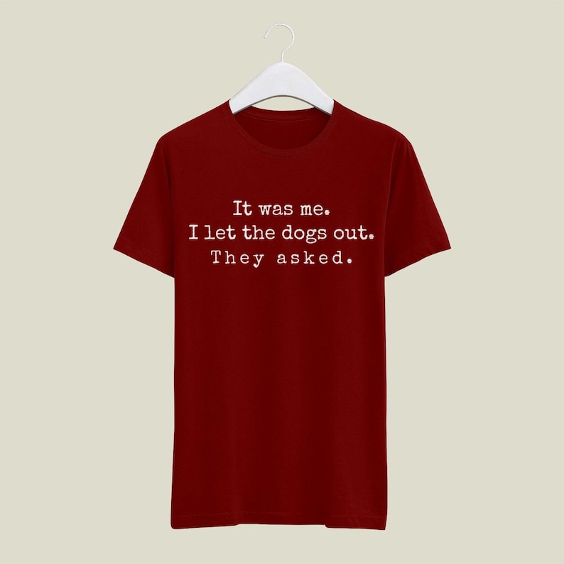 May include: A maroon t-shirt with the text "It was me. I let the dogs out. They asked." in white. The shirt is hanging on a white hanger against a neutral background. The text is centered on the front of the shirt.