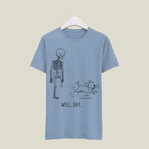 Puede incluir: Una camiseta azul claro con un dibujo lineal negro de un esqueleto al que le falta una pierna y un perro que sale corriendo con un hueso en la boca. Debajo de la ilustraci&oacute;n, el texto "WELL, SHIT..." aparece en una fuente negra sencilla.