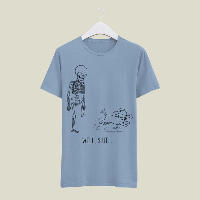 Puede incluir: Una camiseta azul claro con un dibujo lineal negro de un esqueleto al que le falta una pierna y un perro que sale corriendo con un hueso en la boca. Debajo de la ilustraci&oacute;n, el texto "WELL, SHIT..." aparece en una fuente negra sencilla.