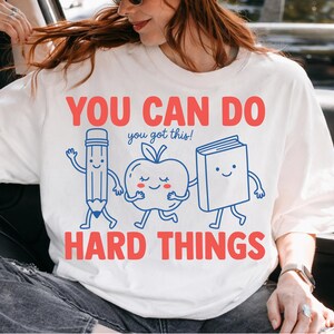 May include: A white short-sleeved t-shirt featuring a graphic print with the red text YOU CAN DO HARD THINGS and the blue text you got this! The design includes cartoon illustrations of a pencil, an apple, and a book with smiling faces.
