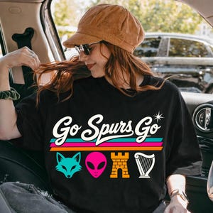 K&ouml;nnte beinhalten: Schwarzes T-Shirt mit dem Text "Go Spurs Go" in Retro-Schrift, mit einem t&uuml;rkisfarbenen Fuchs, einem rosa Alien, einer Backsteinburg und einer wei&szlig;en Harfengrafik. Das T-Shirt wird von einer Person in einem Auto getragen.