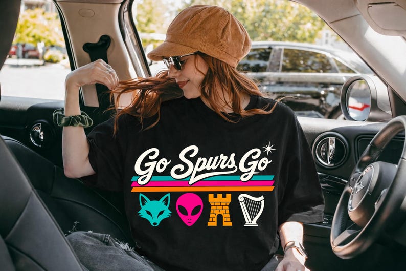 K&ouml;nnte beinhalten: Schwarzes T-Shirt mit dem Text "Go Spurs Go" in Retro-Schrift, mit einem t&uuml;rkisfarbenen Fuchs, einem rosa Alien, einer Backsteinburg und einer wei&szlig;en Harfengrafik. Das T-Shirt wird von einer Person in einem Auto getragen.