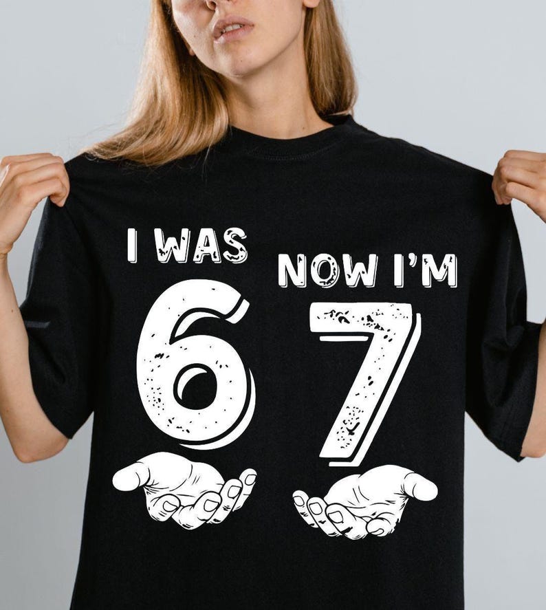 May include: Black t-shirt with the text "I WAS 67 NOW I'M" in white. The numbers 6 and 7 are large and white, with a distressed texture. The number 6 is above a pair of open hands.