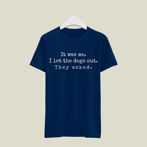 May include: Navy blue t-shirt with the text "It was me. I let the dogs out. They asked." in white, displayed in a simple, sans-serif font. The shirt is hanging on a white hanger.