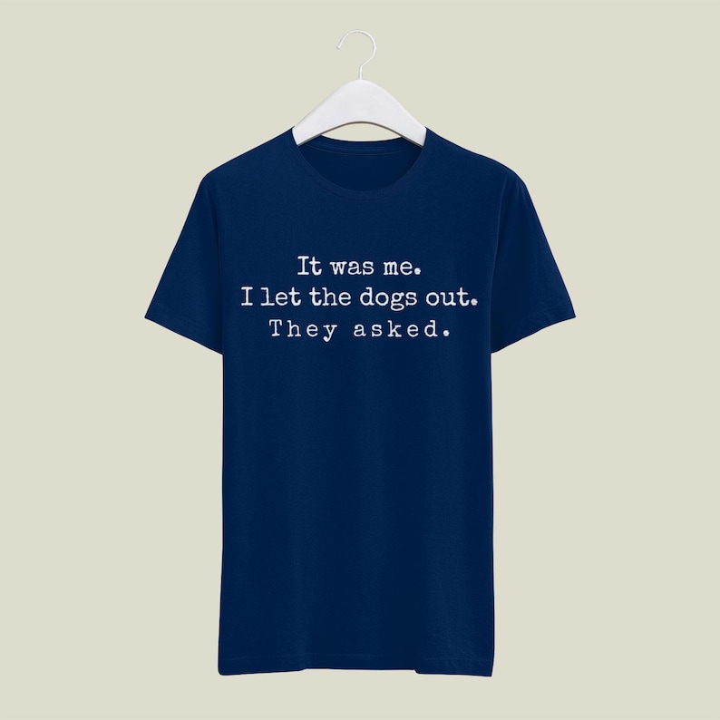 May include: Navy blue t-shirt with the text "It was me. I let the dogs out. They asked." in white, displayed in a simple, sans-serif font. The shirt is hanging on a white hanger.