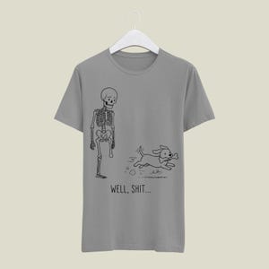 Puede incluir: Una camiseta gris de manga corta con un dibujo lineal negro de un esqueleto al que le falta una pierna y un perro que huye con un hueso en la boca. Debajo de la ilustraci&oacute;n, el texto "WELL, SHIT..." aparece en una fuente negra sencilla.