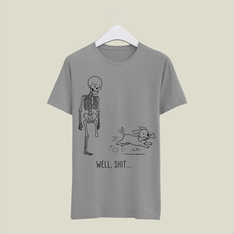 Puede incluir: Una camiseta gris de manga corta con un dibujo lineal negro de un esqueleto al que le falta una pierna y un perro que huye con un hueso en la boca. Debajo de la ilustraci&oacute;n, el texto "WELL, SHIT..." aparece en una fuente negra sencilla.