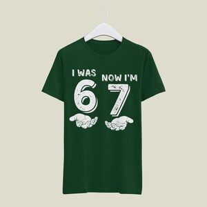 May include: A dark green t-shirt with the text "I WAS NOW I'M 67" in white, with a distressed font. The number 67 is centered, with stylized hands below. The shirt is hanging on a white hanger.