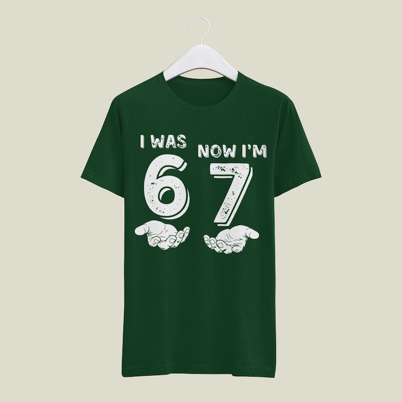 May include: A dark green t-shirt with the text "I WAS NOW I'M 67" in white, with a distressed font. The number 67 is centered, with stylized hands below. The shirt is hanging on a white hanger.