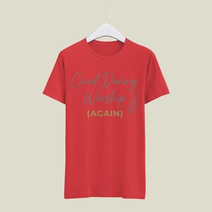May include: A red t-shirt with the text "Cried During Worship (AGAIN)" in a light green script. The shirt is hanging on a white hanger against a neutral background. The design includes a small floral element.