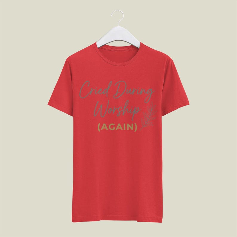 May include: A red t-shirt with the text "Cried During Worship (AGAIN)" in a light green script. The shirt is hanging on a white hanger against a neutral background. The design includes a small floral element.