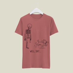 Puede incluir: Una camiseta color rosa empolvado con un gr&aacute;fico de l&iacute;neas negras de un esqueleto al que le falta una pierna y un perro que huye con un hueso en la boca. Debajo de la ilustraci&oacute;n, el texto dice "WELL, SHIT..." en una fuente negra sencilla.