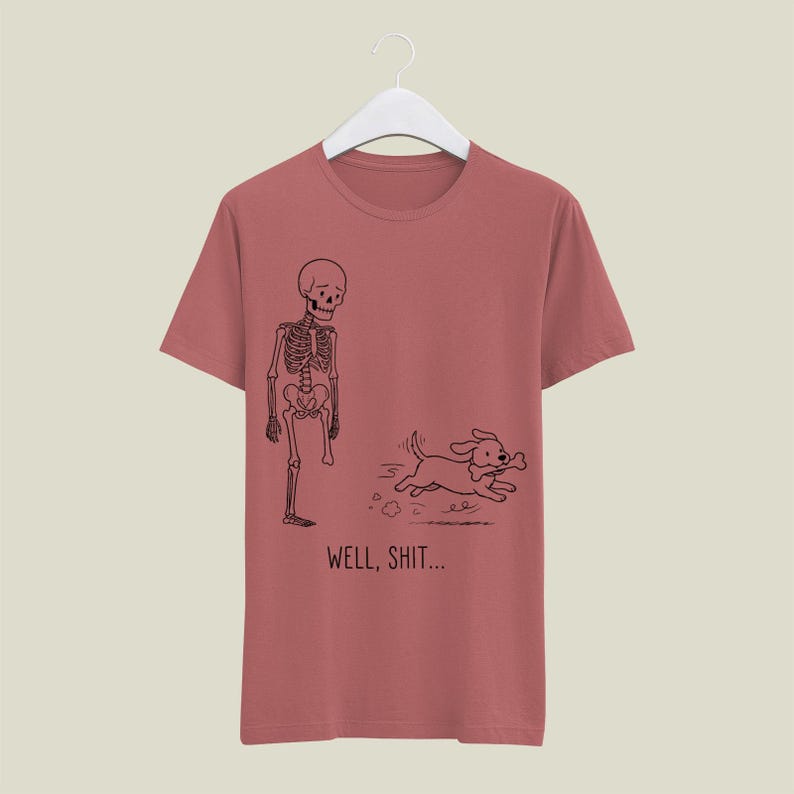 Puede incluir: Una camiseta color rosa empolvado con un gr&aacute;fico de l&iacute;neas negras de un esqueleto al que le falta una pierna y un perro que huye con un hueso en la boca. Debajo de la ilustraci&oacute;n, el texto dice "WELL, SHIT..." en una fuente negra sencilla.